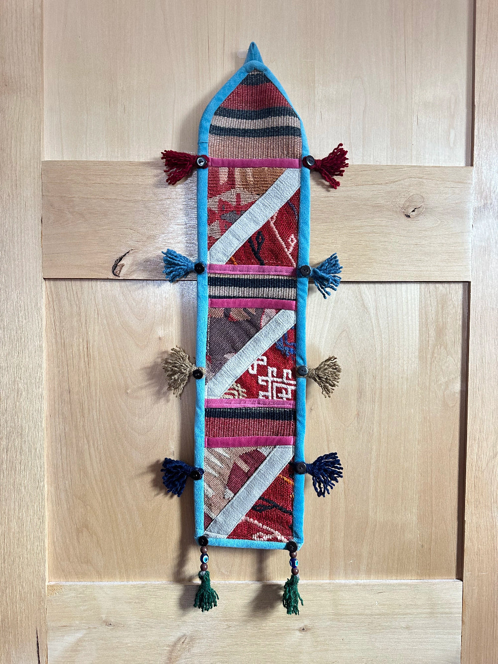 Handmade Turkish wall hanging with vintage rug patterns, featuring colorful tassels, evil eye beads, and geometric designs on a wood panel background. Unique bohemian decor made from authentic Turkish rug fragments, perfect for home styling.