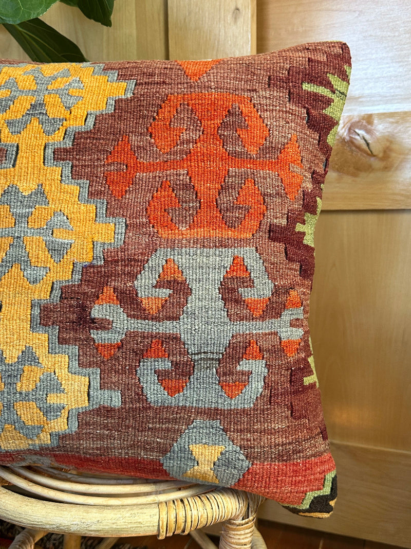 Square vintage Turkish kilim pillow with bold geometric patterns in yellow, orange, and light blue on a deep red wool background, handwoven from repurposed rug fragments.