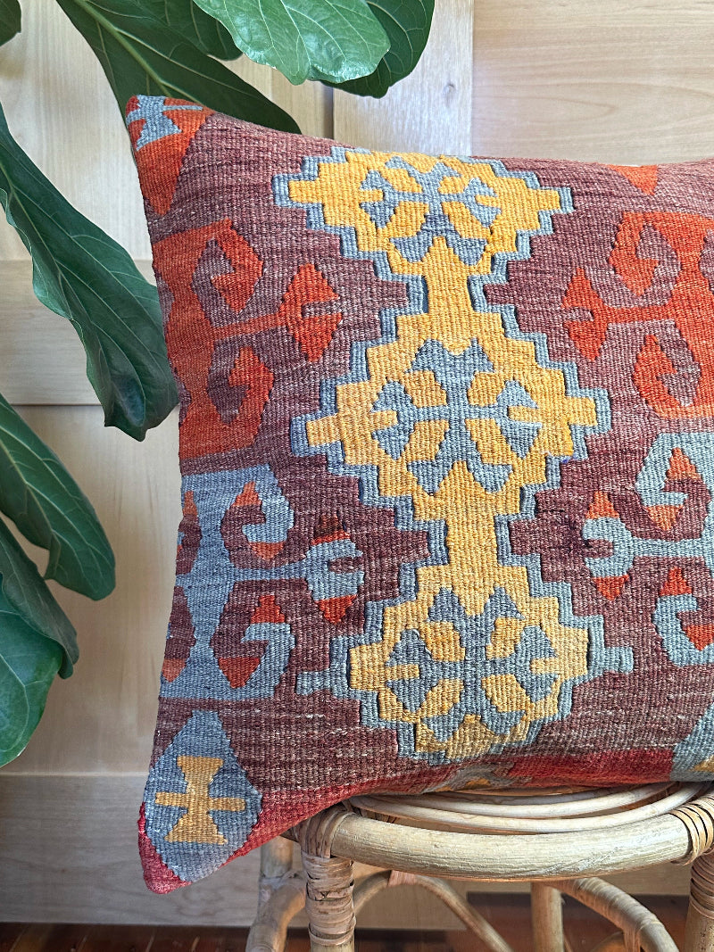 Square vintage Turkish kilim pillow with bold geometric patterns in yellow, orange, and light blue on a deep red wool background, handwoven from repurposed rug fragments.
