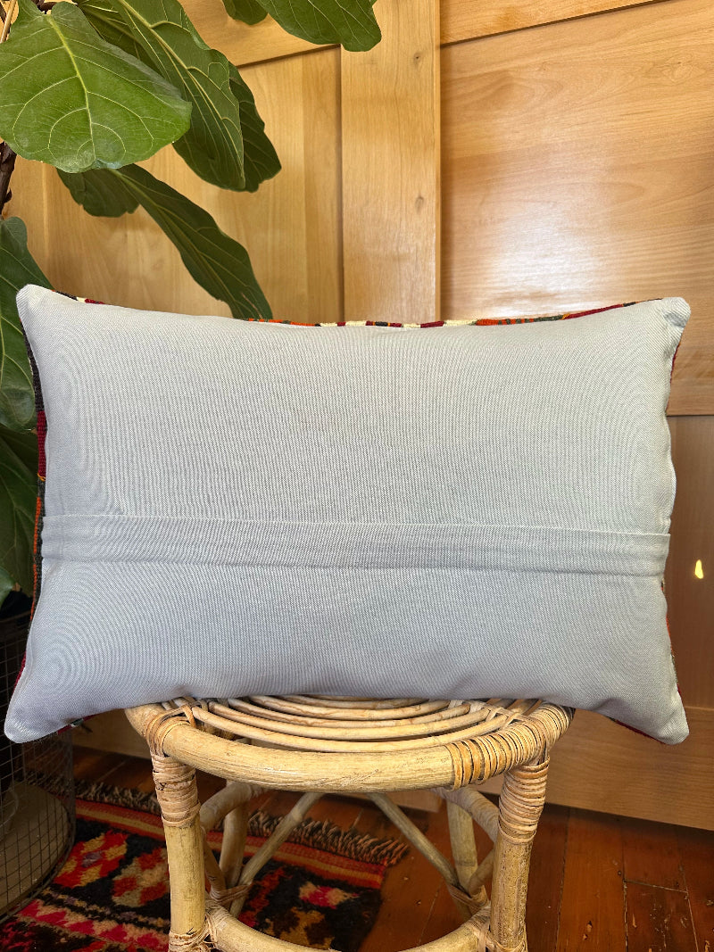Gray pillow on a wicker stool with a wooden wall and plant in the background