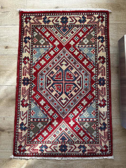 Handknotted two by three foot mini Kazak rug featuring intricate geometric patterns in rich red, deep blue, and warm neutral tones. Made from 100% wool with a durable low-pile construction, perfect for high-traffic areas. Ideal for vintage, traditional, and eclectic home décor.