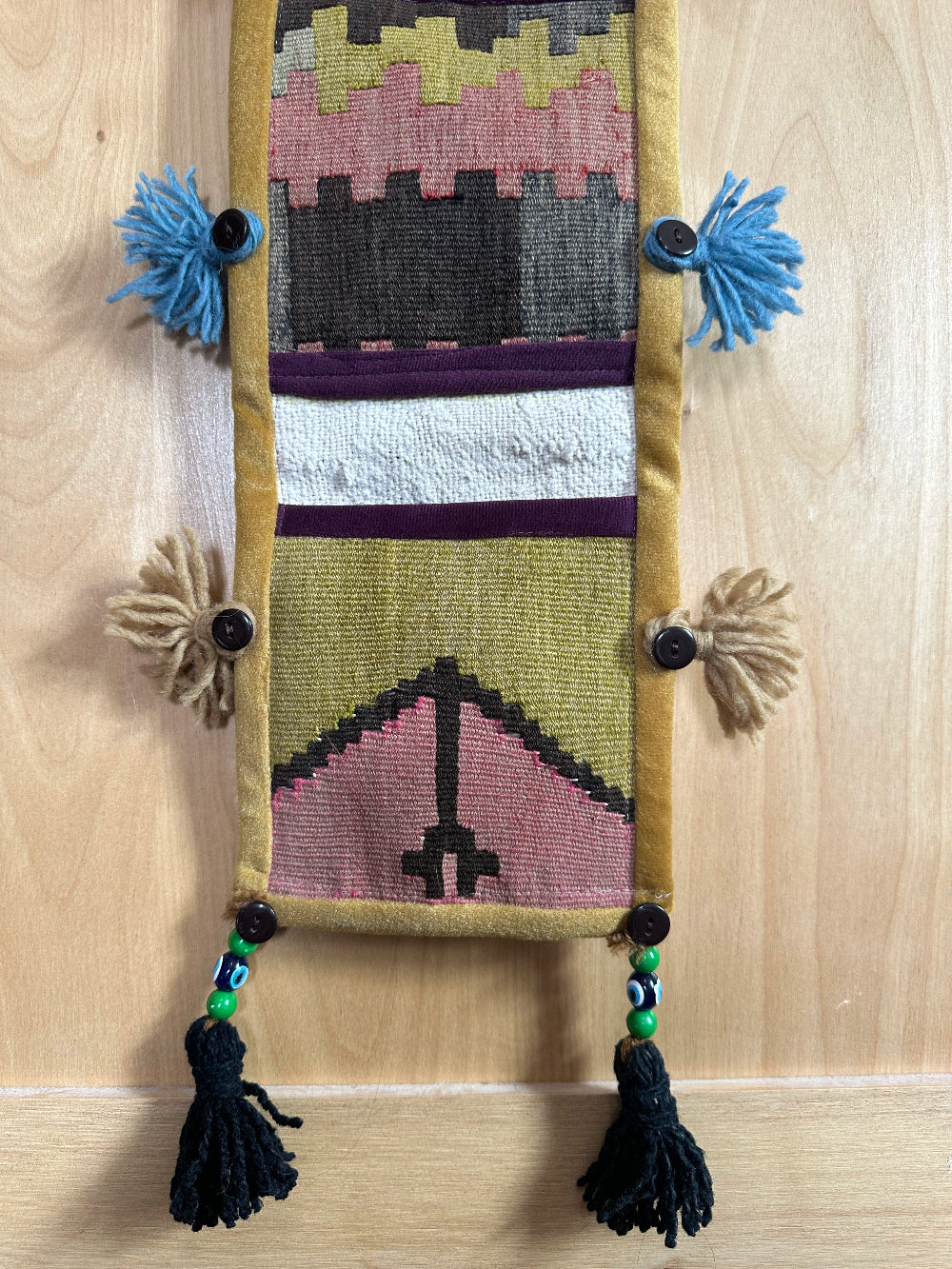 Handmade Turkish wall hanging with vintage rug patterns, featuring colorful tassels, evil eye beads, and geometric designs on a wood panel background. Unique bohemian decor made from authentic Turkish rug fragments, perfect for home styling.