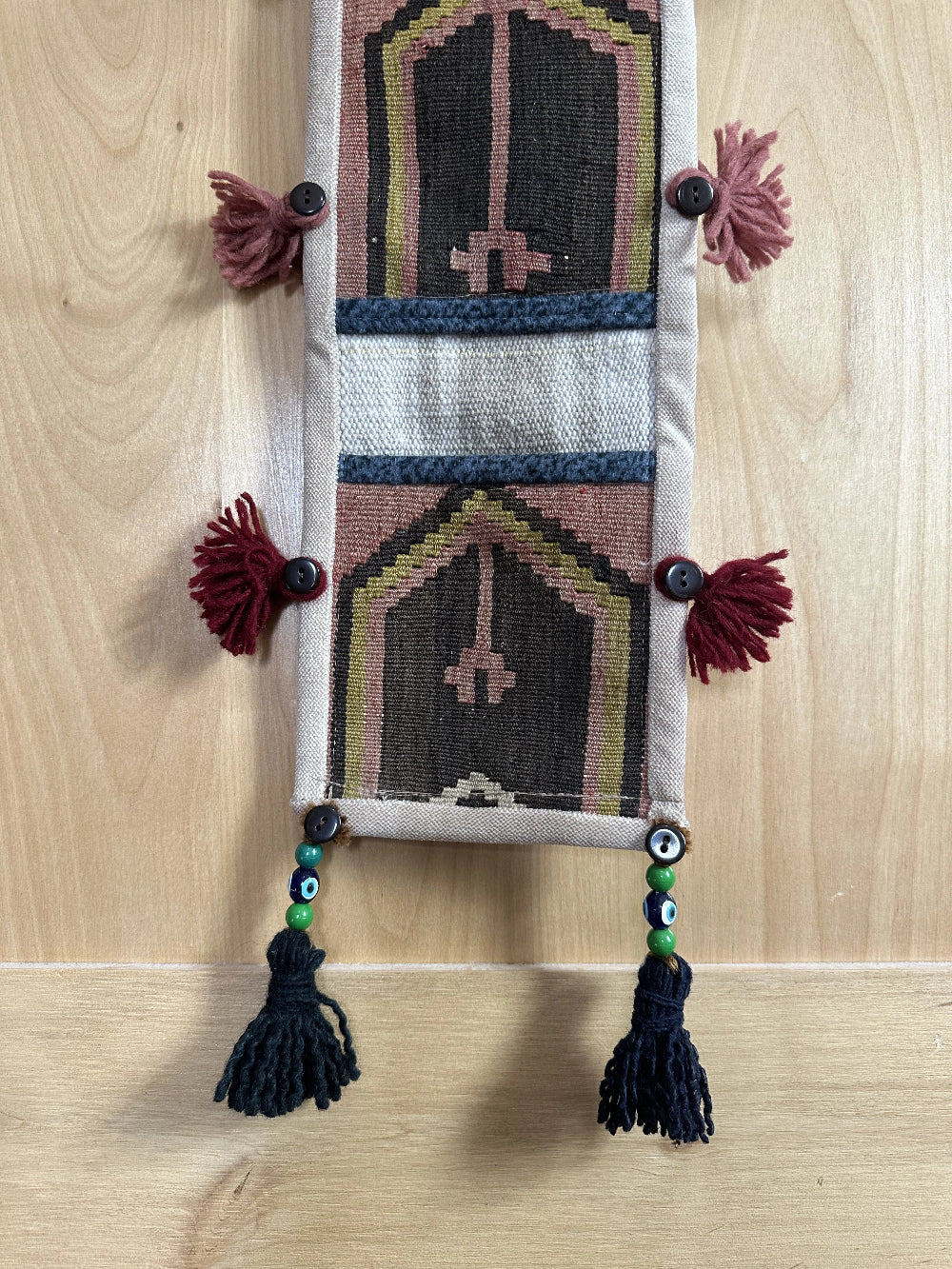 Handmade Turkish wall hanging with vintage rug patterns, featuring colorful tassels, evil eye beads, and geometric designs on a wood panel background. Unique bohemian decor made from authentic Turkish rug fragments, perfect for home styling.