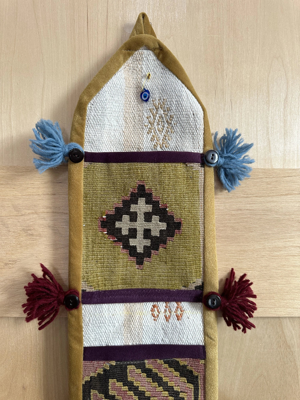 Handmade Turkish wall hanging with vintage rug patterns, featuring colorful tassels, evil eye beads, and geometric designs on a wood panel background. Unique bohemian decor made from authentic Turkish rug fragments, perfect for home styling.