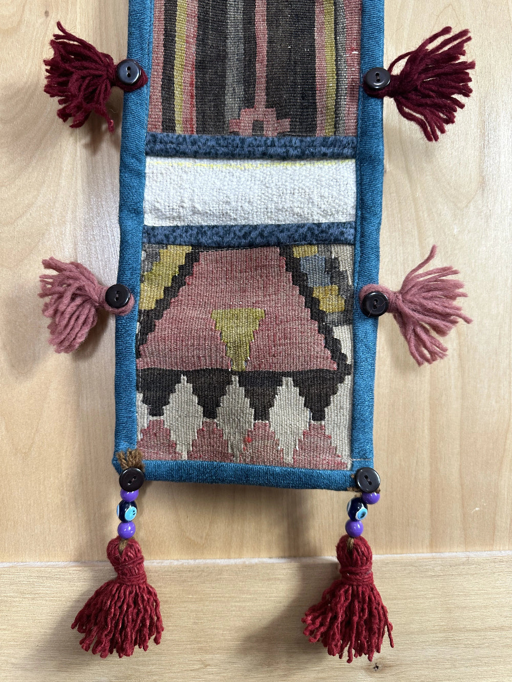 Handmade Turkish wall hanging with vintage rug patterns, featuring colorful tassels, evil eye beads, and geometric designs on a wood panel background. Unique bohemian decor made from authentic Turkish rug fragments, perfect for home styling.