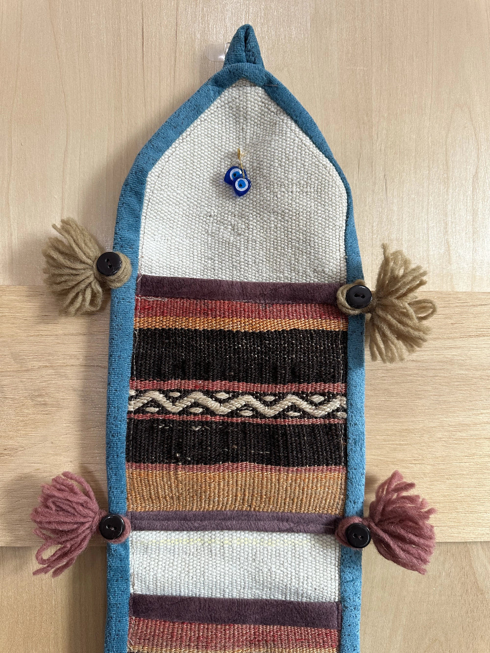 Handmade Turkish wall hanging with vintage rug patterns, featuring colorful tassels, evil eye beads, and geometric designs on a wood panel background. Unique bohemian decor made from authentic Turkish rug fragments, perfect for home styling.