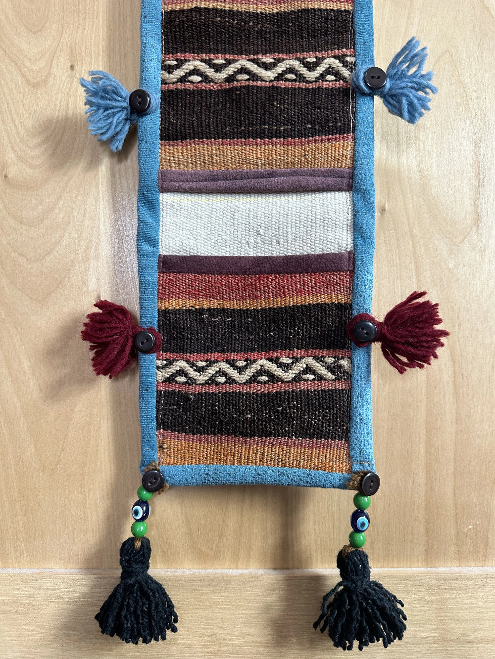 Handmade Turkish wall hanging with vintage rug patterns, featuring colorful tassels, evil eye beads, and geometric designs on a wood panel background. Unique bohemian decor made from authentic Turkish rug fragments, perfect for home styling.