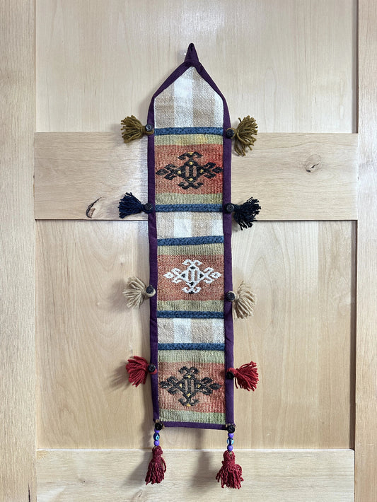 Handmade Turkish wall hanging with vintage rug patterns, featuring colorful tassels, evil eye beads, and geometric designs on a wood panel background. Unique bohemian decor made from authentic Turkish rug fragments, perfect for home styling.