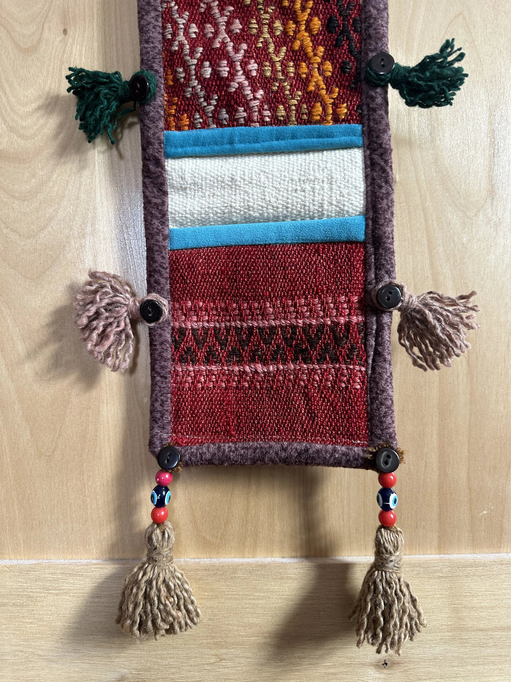 Handmade Turkish wall hanging with vintage rug patterns, featuring colorful tassels, evil eye beads, and geometric designs on a wood panel background. Unique bohemian decor made from authentic Turkish rug fragments, perfect for home styling.