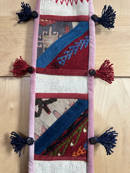 Handmade Turkish wall hanging with vintage rug patterns, featuring colorful tassels, evil eye beads, and geometric designs on a wood panel background. Unique bohemian decor made from authentic Turkish rug fragments, perfect for home styling.