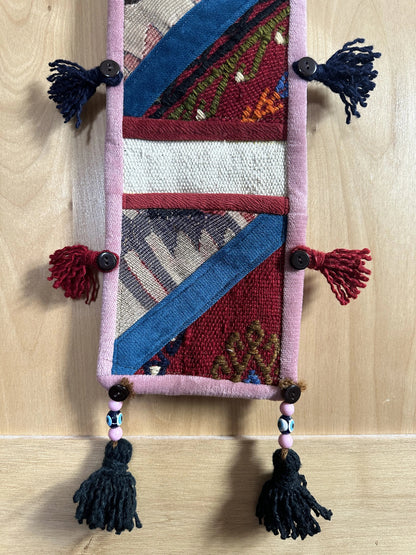 Handmade Turkish wall hanging with vintage rug patterns, featuring colorful tassels, evil eye beads, and geometric designs on a wood panel background. Unique bohemian decor made from authentic Turkish rug fragments, perfect for home styling.