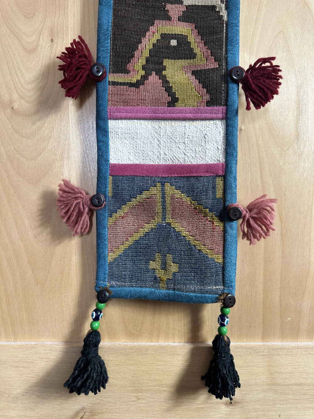 Handmade Turkish wall hanging with vintage rug patterns, featuring colorful tassels, evil eye beads, and geometric designs on a wood panel background. Unique bohemian decor made from authentic Turkish rug fragments, perfect for home styling.