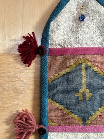 Handmade Turkish wall hanging with vintage rug patterns, featuring colorful tassels, evil eye beads, and geometric designs on a wood panel background. Unique bohemian decor made from authentic Turkish rug fragments, perfect for home styling.