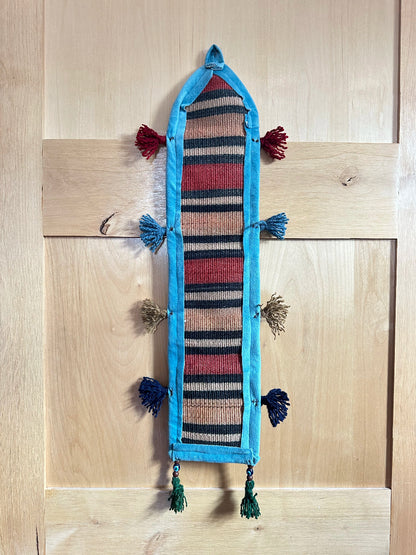 Handmade Turkish wall hanging with vintage rug patterns, featuring colorful tassels, evil eye beads, and geometric designs on a wood panel background. Unique bohemian decor made from authentic Turkish rug fragments, perfect for home styling.