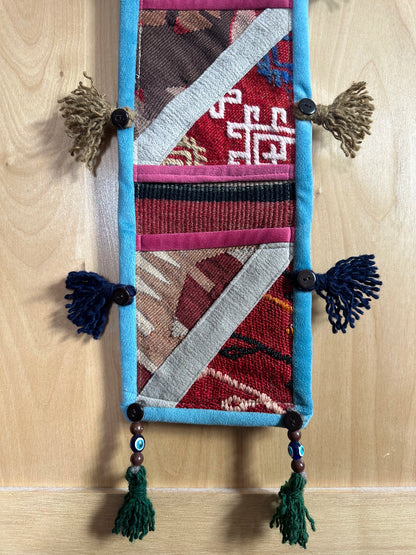 Handmade Turkish wall hanging with vintage rug patterns, featuring colorful tassels, evil eye beads, and geometric designs on a wood panel background. Unique bohemian decor made from authentic Turkish rug fragments, perfect for home styling.