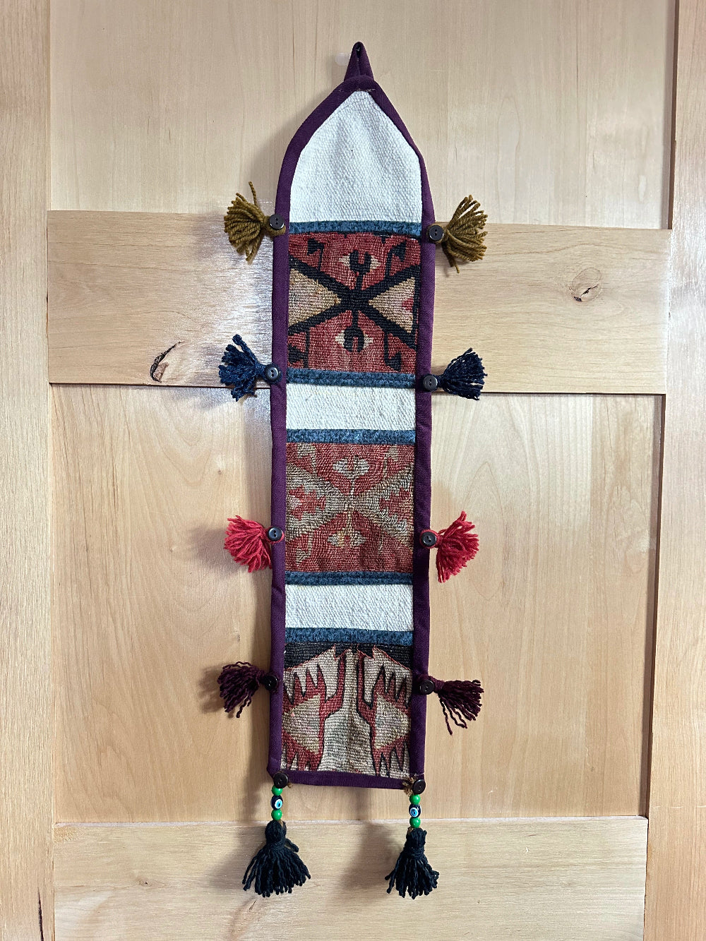 Handmade Turkish wall hanging with vintage rug patterns, featuring colorful tassels, evil eye beads, and geometric designs on a wood panel background. Unique bohemian decor made from authentic Turkish rug fragments, perfect for home styling.