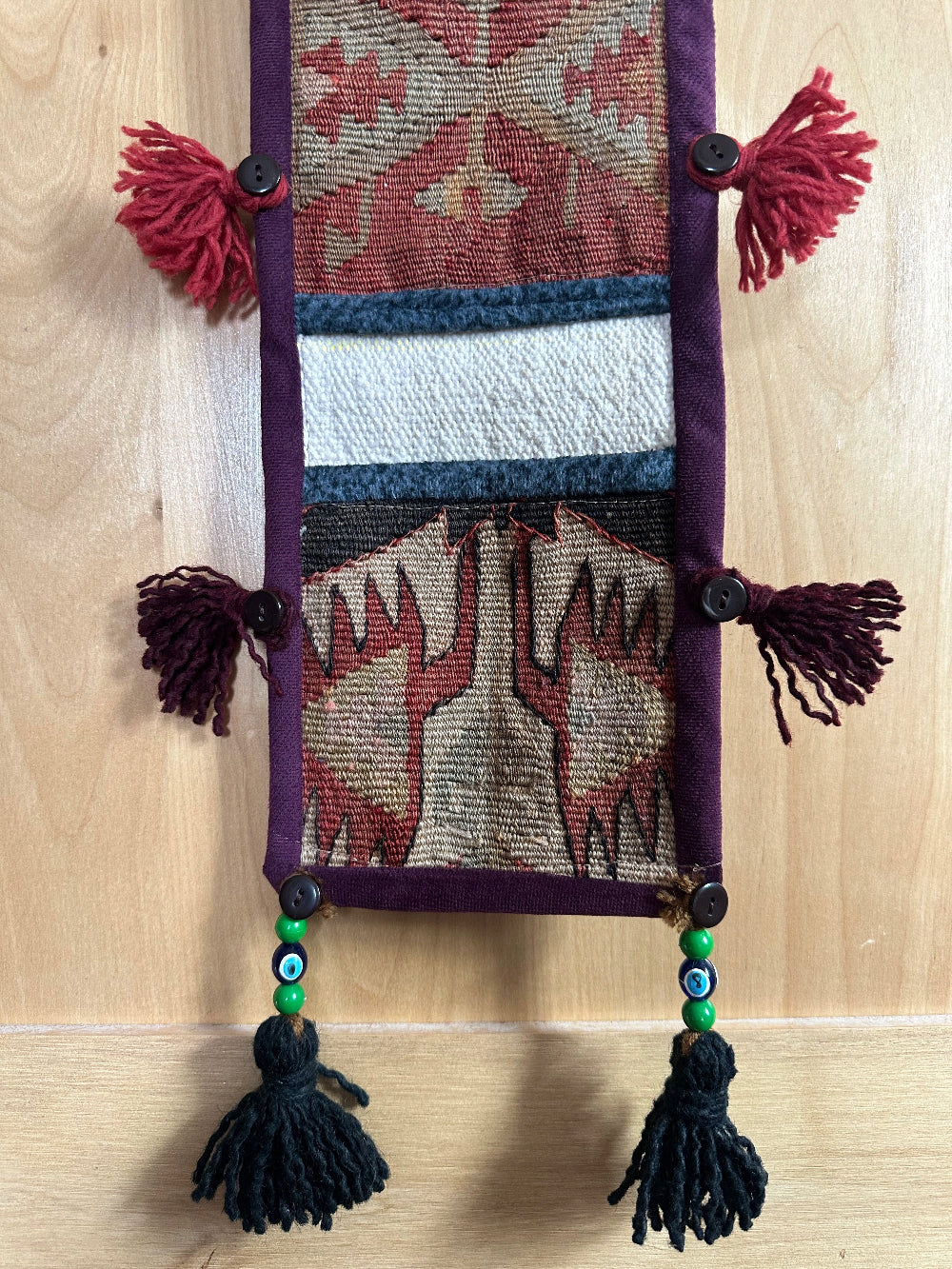 Handmade Turkish wall hanging with vintage rug patterns, featuring colorful tassels, evil eye beads, and geometric designs on a wood panel background. Unique bohemian decor made from authentic Turkish rug fragments, perfect for home styling.