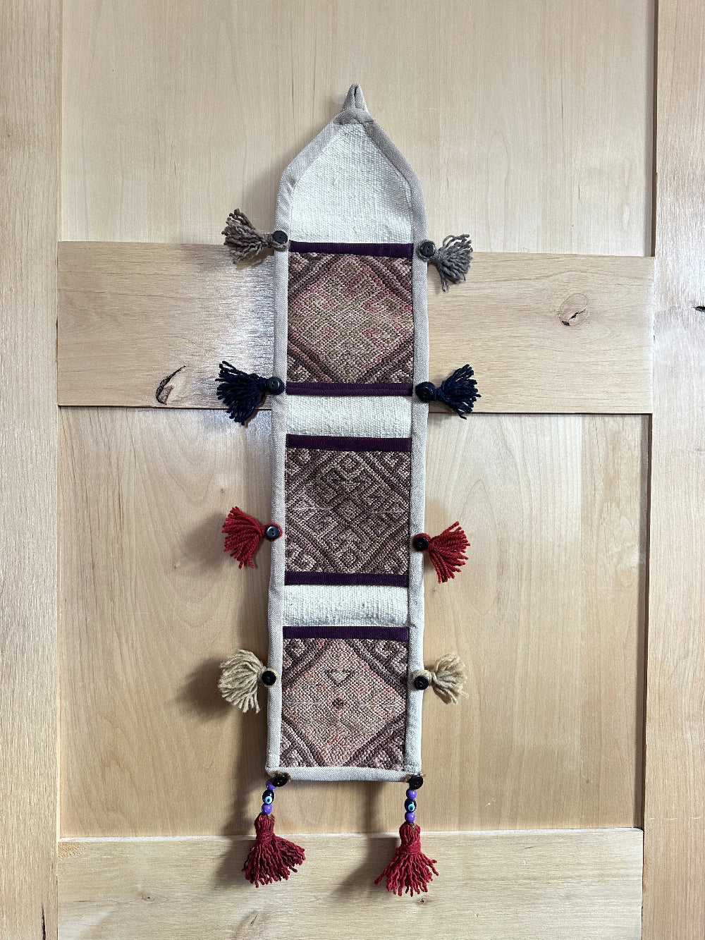 Handmade Turkish wall hanging with vintage rug patterns, featuring colorful tassels, evil eye beads, and geometric designs on a wood panel background. Unique bohemian decor made from authentic Turkish rug fragments, perfect for home styling.