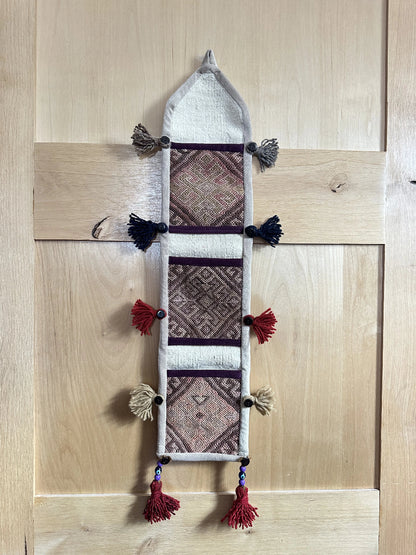 Handmade Turkish wall hanging with vintage rug patterns, featuring colorful tassels, evil eye beads, and geometric designs on a wood panel background. Unique bohemian decor made from authentic Turkish rug fragments, perfect for home styling.