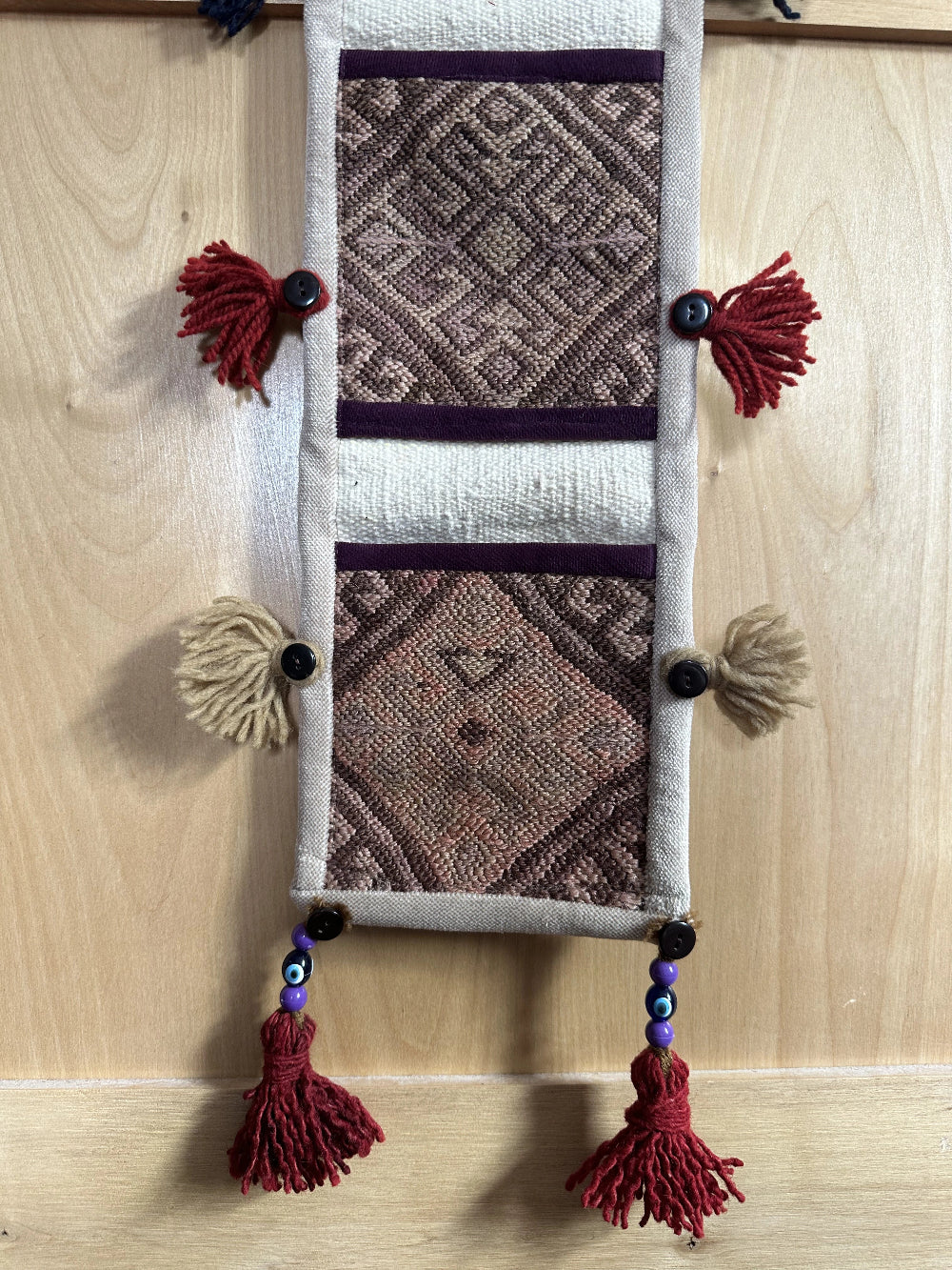 Handmade Turkish wall hanging with vintage rug patterns, featuring colorful tassels, evil eye beads, and geometric designs on a wood panel background. Unique bohemian decor made from authentic Turkish rug fragments, perfect for home styling.