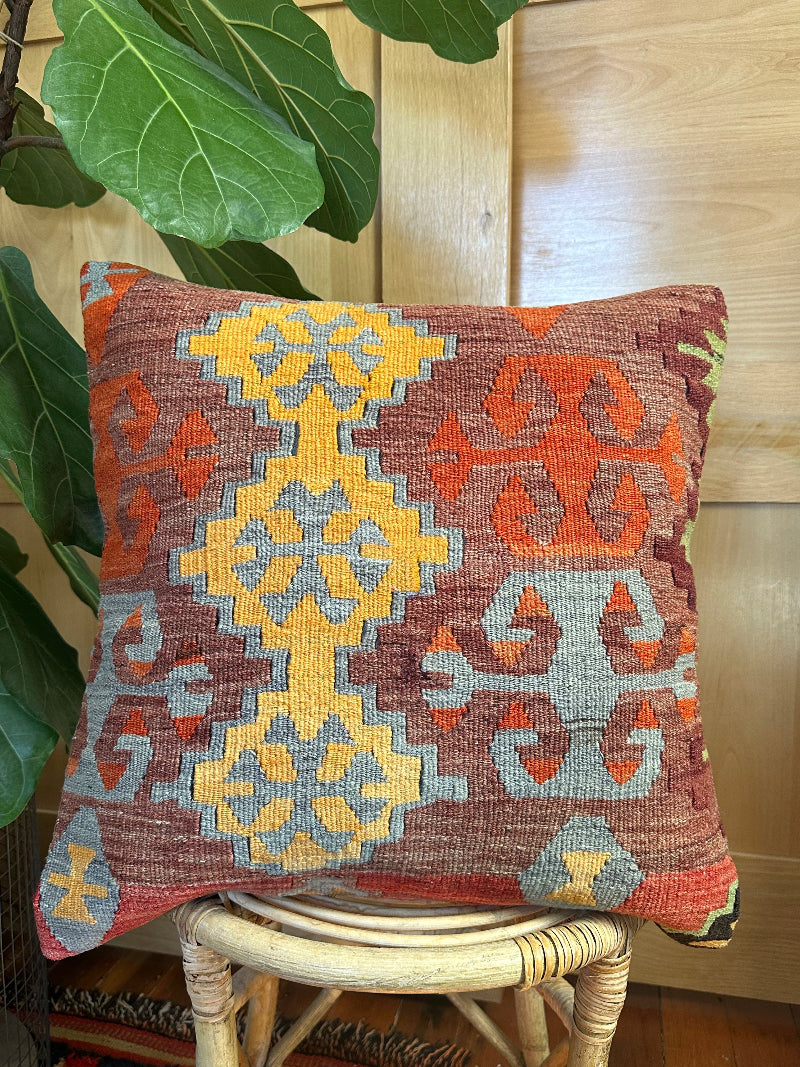 Square vintage Turkish kilim pillow with bold geometric patterns in yellow, orange, and light blue on a deep red wool background, handwoven from repurposed rug fragments.