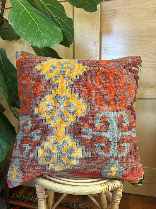 Square vintage Turkish kilim pillow with bold geometric patterns in yellow, orange, and light blue on a deep red wool background, handwoven from repurposed rug fragments.