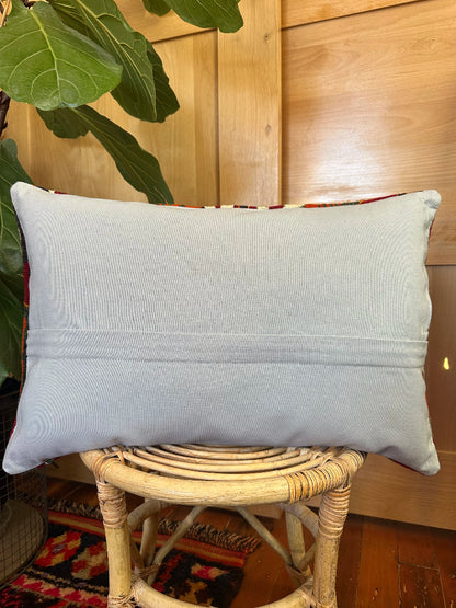 Gray pillow on a wicker stool with a wooden wall and plant in the background