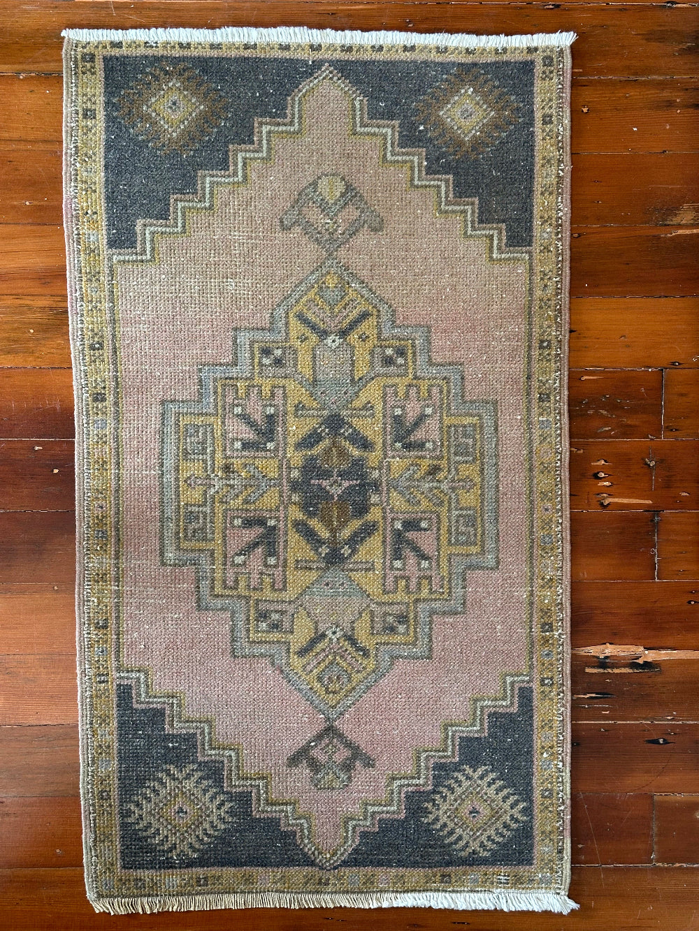 Vintage Turkish wool rug with a central geometric medallion in muted pink, gold, and dark charcoal tones, featuring stepped corner motifs and a distressed finish.