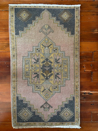 Vintage Turkish wool rug with a central geometric medallion in muted pink, gold, and dark charcoal tones, featuring stepped corner motifs and a distressed finish.