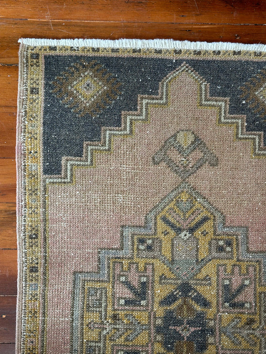 Vintage Turkish wool rug with a central geometric medallion in muted pink, gold, and dark charcoal tones, featuring stepped corner motifs and a distressed finish.