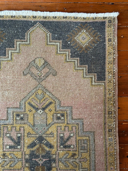 Vintage Turkish wool rug with a central geometric medallion in muted pink, gold, and dark charcoal tones, featuring stepped corner motifs and a distressed finish.