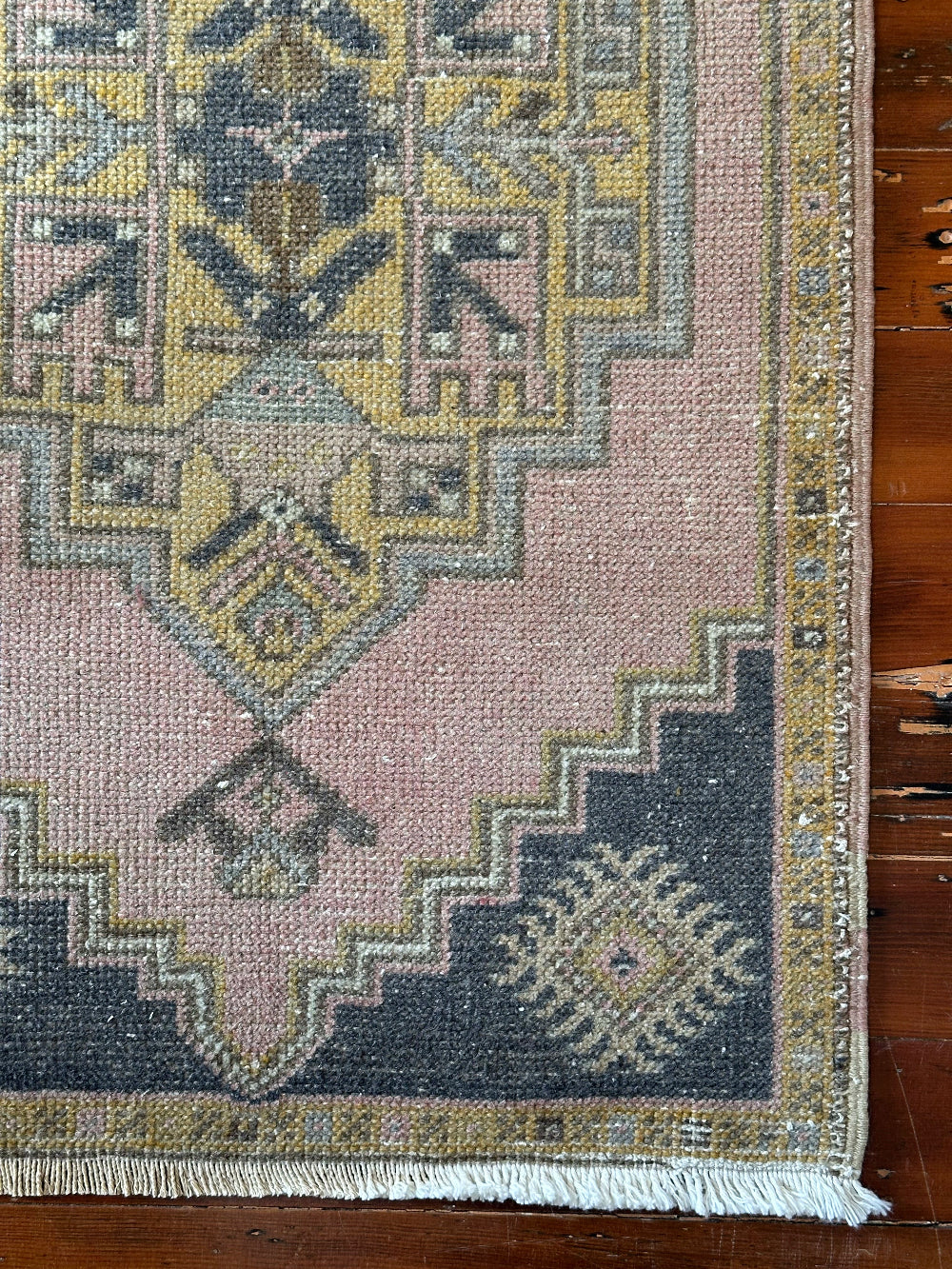 Vintage Turkish wool rug with a central geometric medallion in muted pink, gold, and dark charcoal tones, featuring stepped corner motifs and a distressed finish.