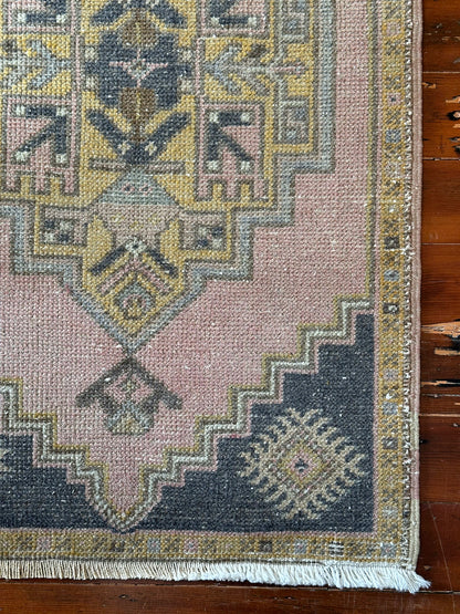Vintage Turkish wool rug with a central geometric medallion in muted pink, gold, and dark charcoal tones, featuring stepped corner motifs and a distressed finish.