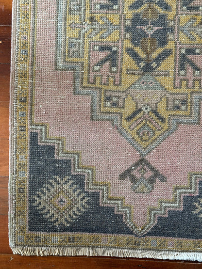 Vintage Turkish wool rug with a central geometric medallion in muted pink, gold, and dark charcoal tones, featuring stepped corner motifs and a distressed finish.