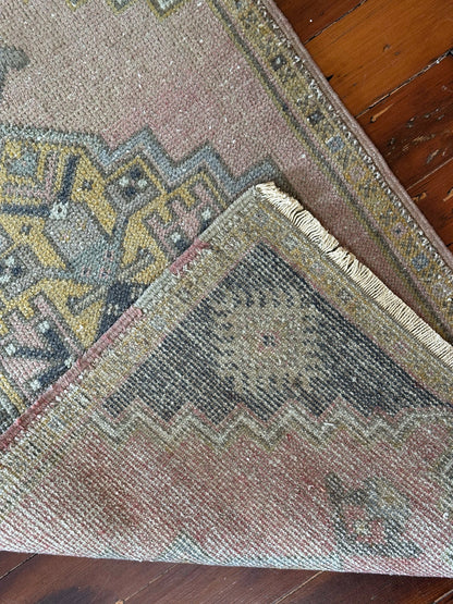 Vintage Turkish wool rug with a central geometric medallion in muted pink, gold, and dark charcoal tones, featuring stepped corner motifs and a distressed finish.