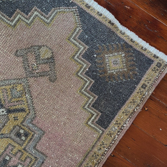 Vintage Turkish wool rug with a central geometric medallion in muted pink, gold, and dark charcoal tones, featuring stepped corner motifs and a distressed finish.